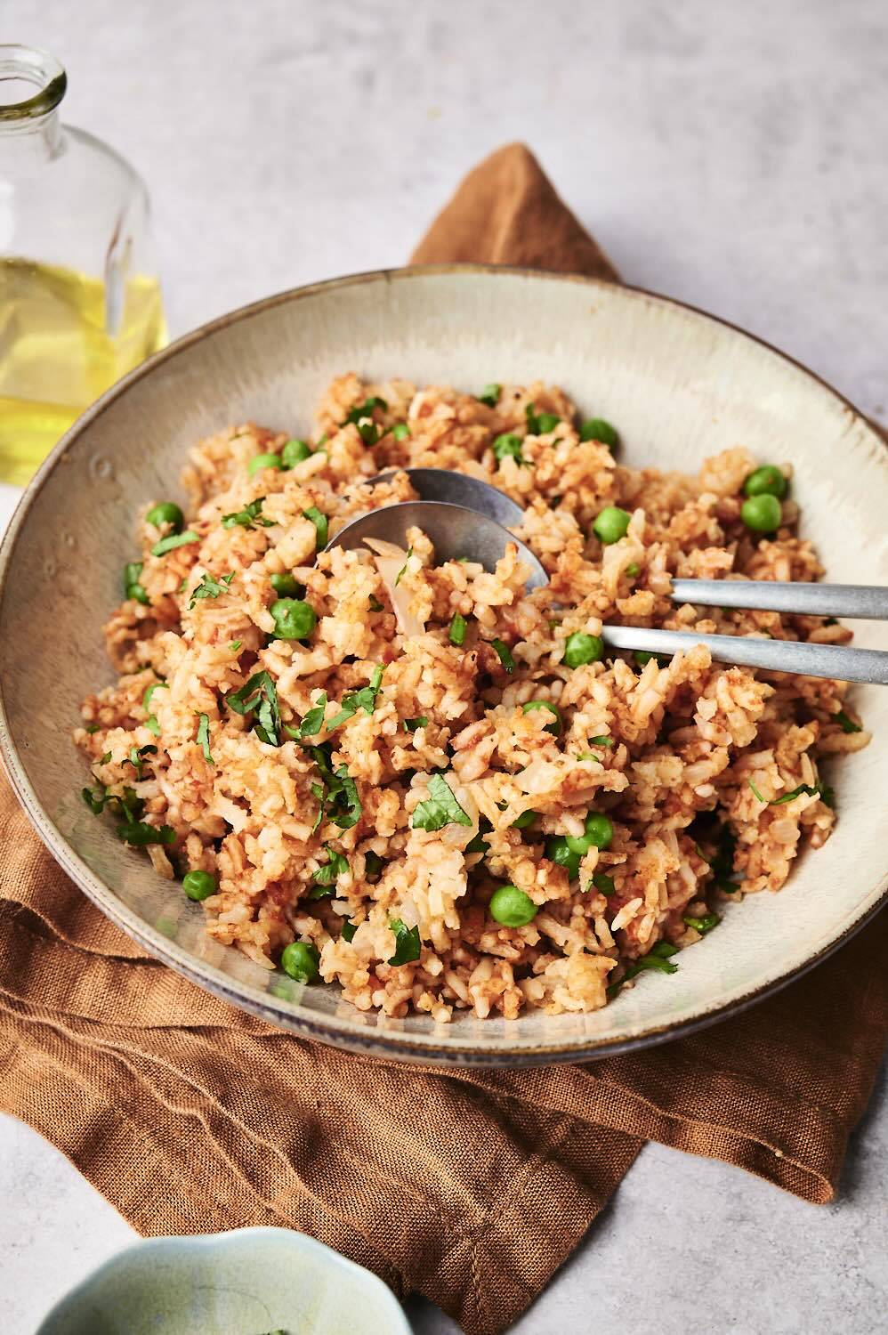 A bowl of fluffy slow cooker Mexican rice with green peas and fresh cilantro, served with silver spoons on a brown napkin. 