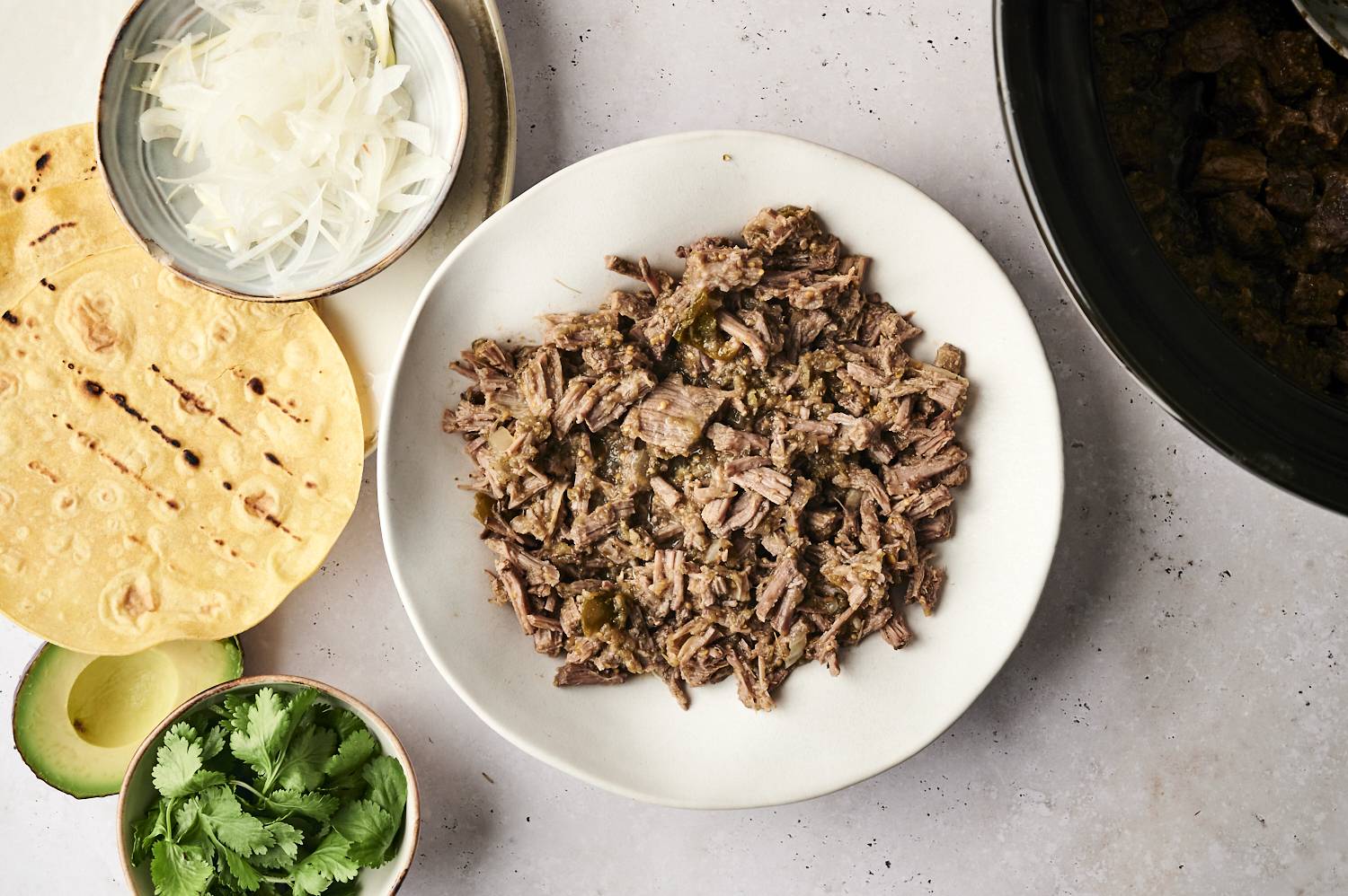 Shredded salsa verde beef served in a white bowl, surrounded by grilled tortillas, sliced onions, fresh cilantro, and avocado, ready for taco assembly.