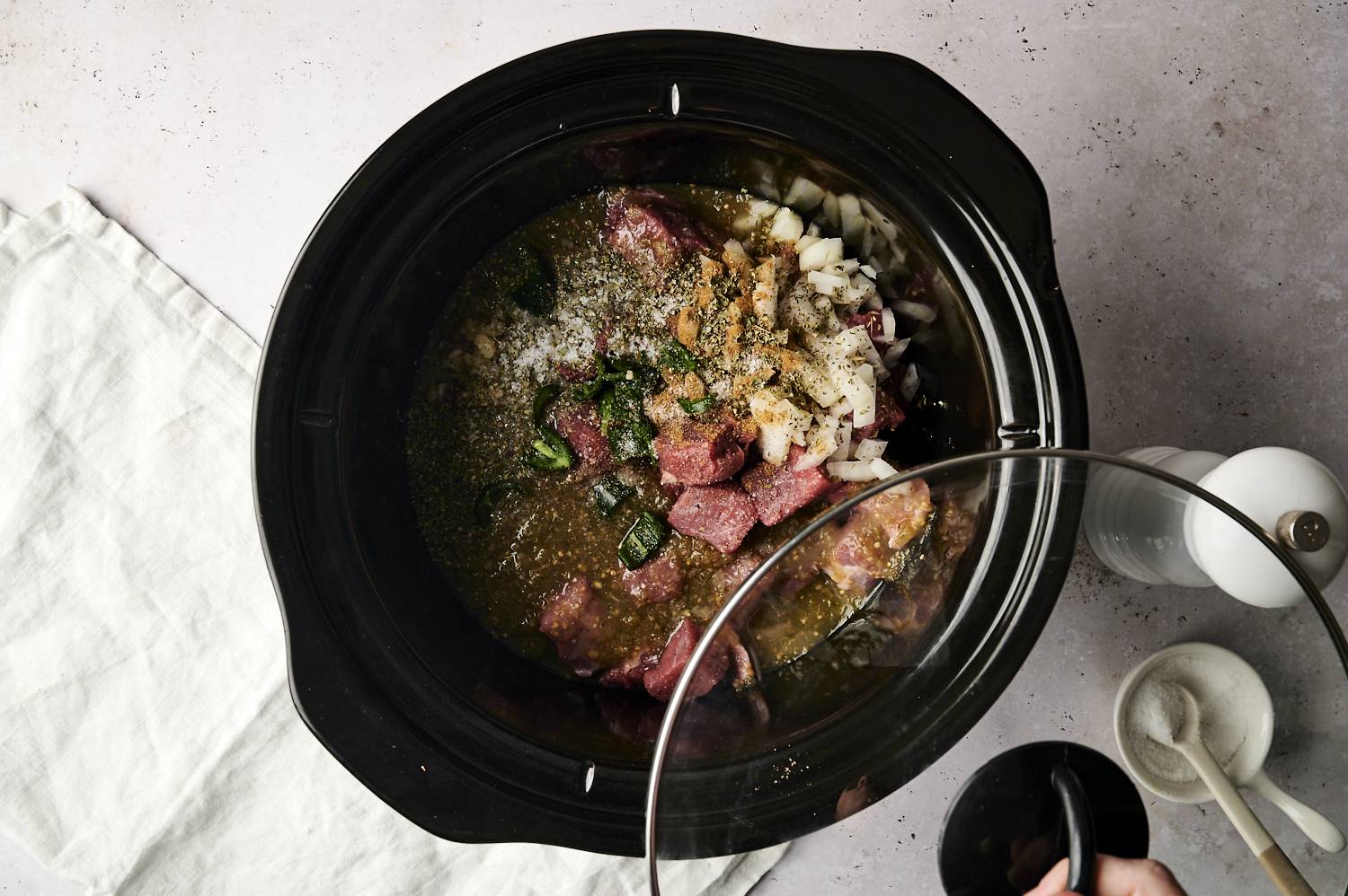 Cubed beef, diced onions, jalapeños, spices, and salsa verde inside a slow cooker, with a hand placing the lid on top, preparing the ingredients for cooking