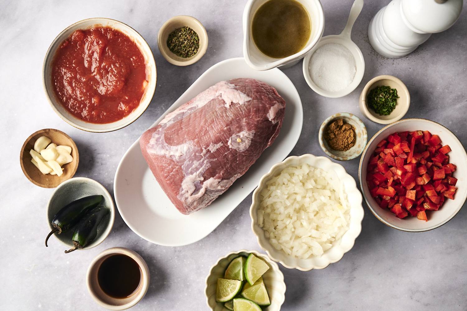 A flat lay of fresh ingredients including a raw beef roast, diced tomatoes, onions, red bell peppers, garlic, jalapeños, spices, lime wedges, and broth, arranged on a gray surface.