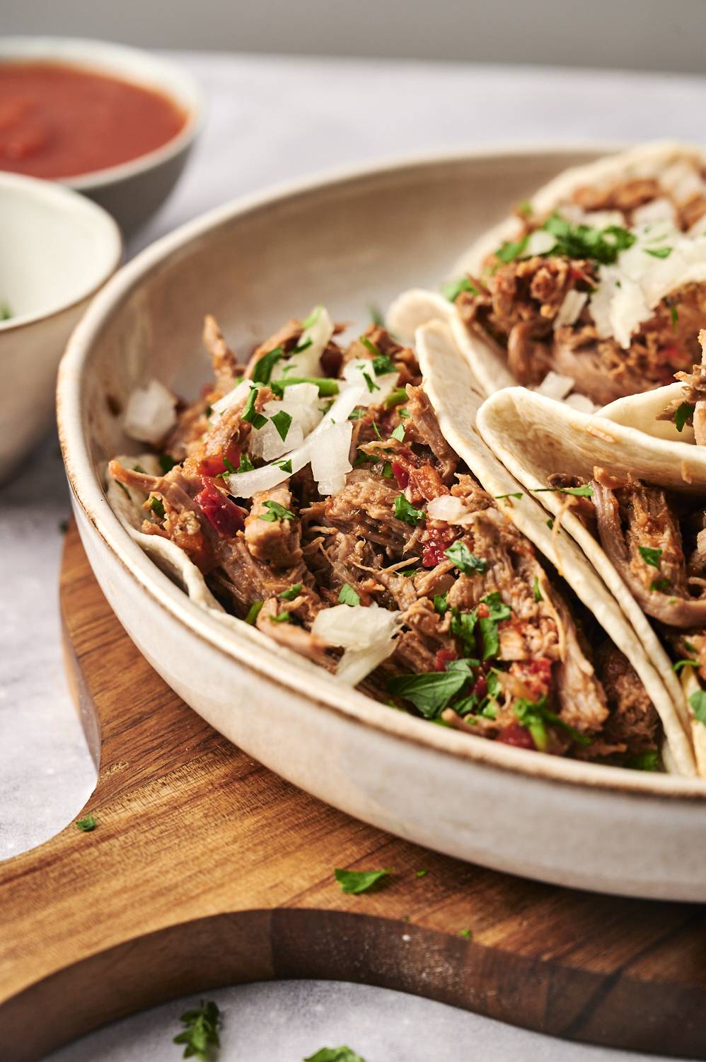 A close-up of slow-cooked spicy braised beef tacos in soft flour tortillas, topped with chopped onions and fresh cilantro, served on a wooden board.