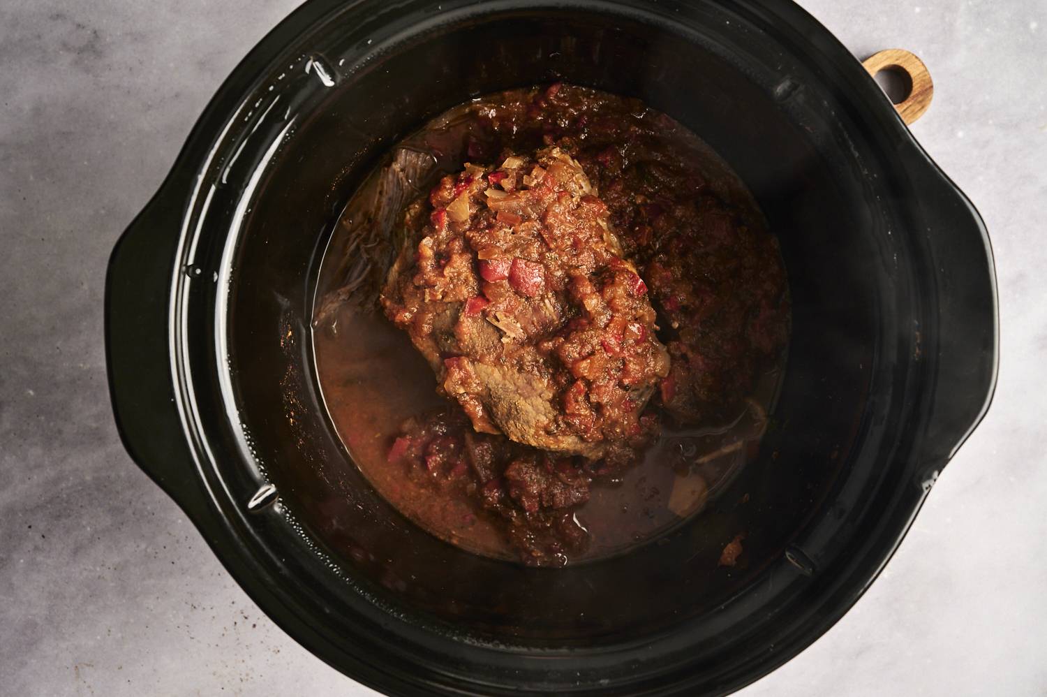 A slow-cooked beef roast in a black slow cooker, covered in a rich, spiced tomato sauce, ready to be shredded.