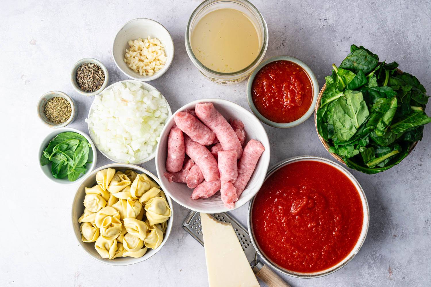 Fresh ingredients for Slow Cooker Dumpling Soup on a flat lay including sausage, onions, garlic, spinach, dumplings, crushed tomatoes, tomato paste, chicken stock, herbs, basil and a cube of parmesan cheese ready to be grated.