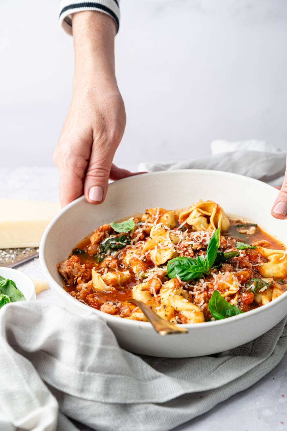 One holds a bowl of Slow Cooker Dumpling Soup, filled with sausage, spinach and cheese dumplings that are rich and saucy and topped with fresh basil and grated Parmesan for a comforting, homemade flavor.