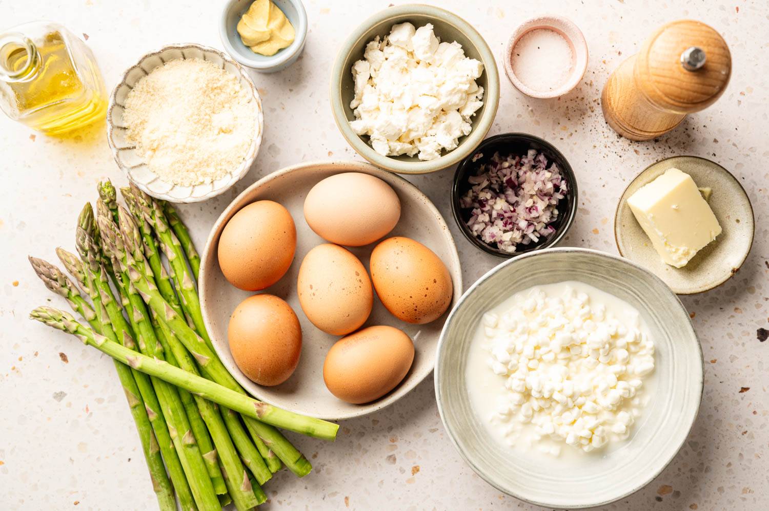 A collection of fresh ingredients on a countertop, including asparagus, six brown eggs, cottage cheese, butter, chopped onions, parmesan, and olive oil.