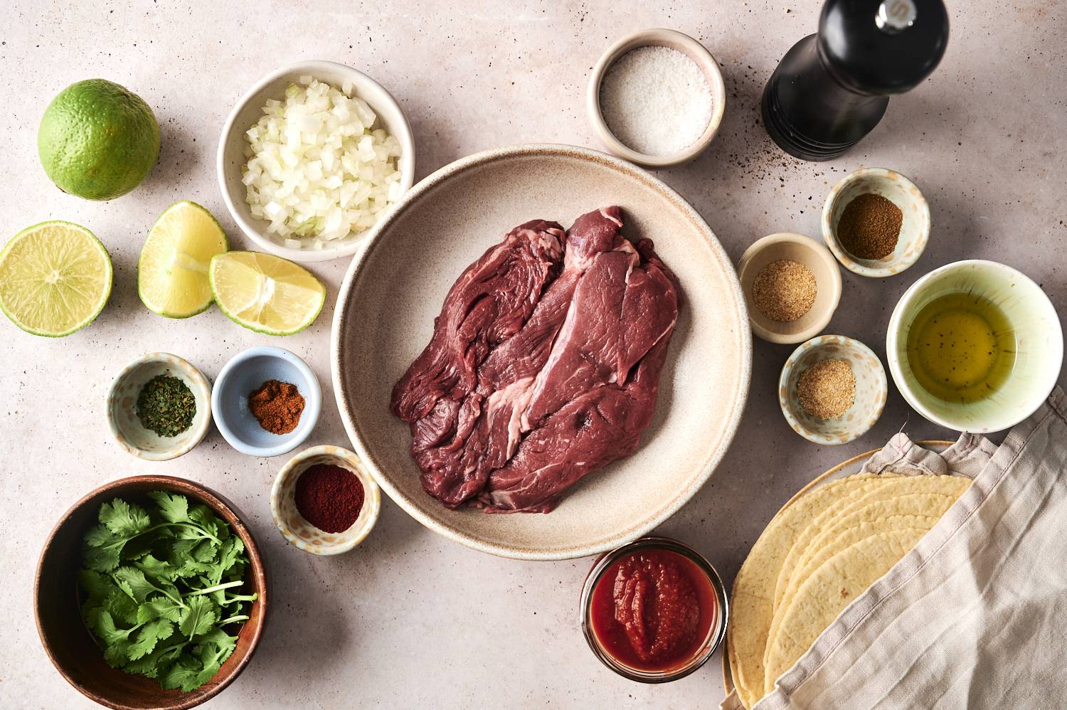 Flat steak taco ingredients including raw steak, seasoning, chopped onion, lemon wedges, salsa, coriander and tortillas.