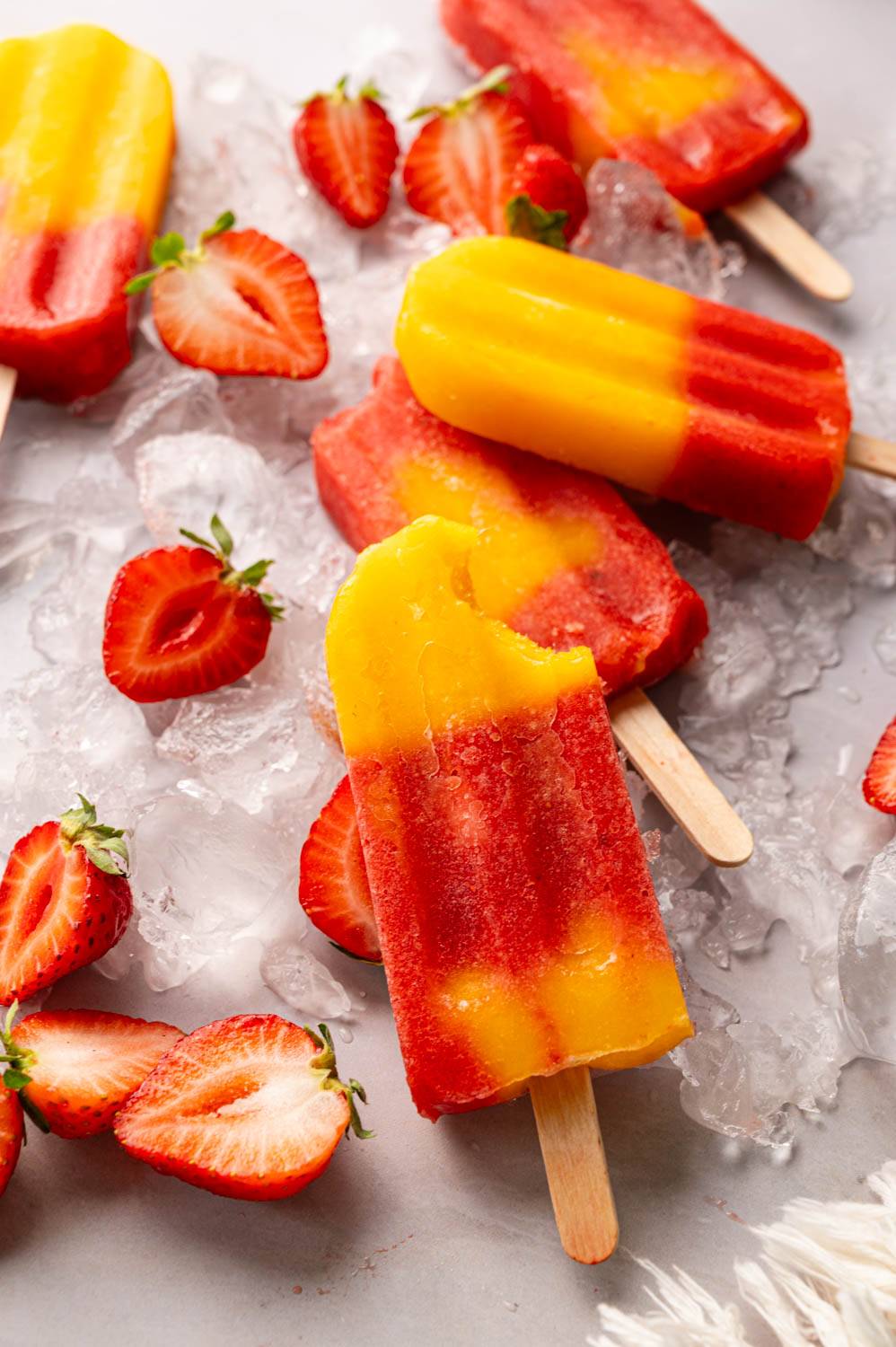 Homemade strawberry mango popsicles with vibrant red and yellow layers served over crushed ice with fresh strawberries.