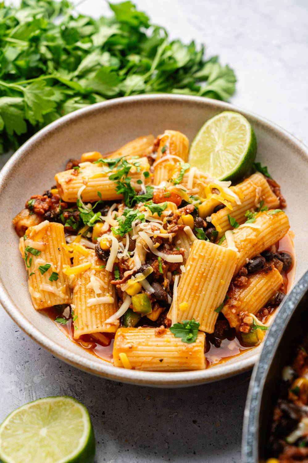 A bowl of macaroni with black beans, corn and tomato sauce served with shredded cheese and cilantro and a lime wedge on the side. Fresh cilantro in the background.