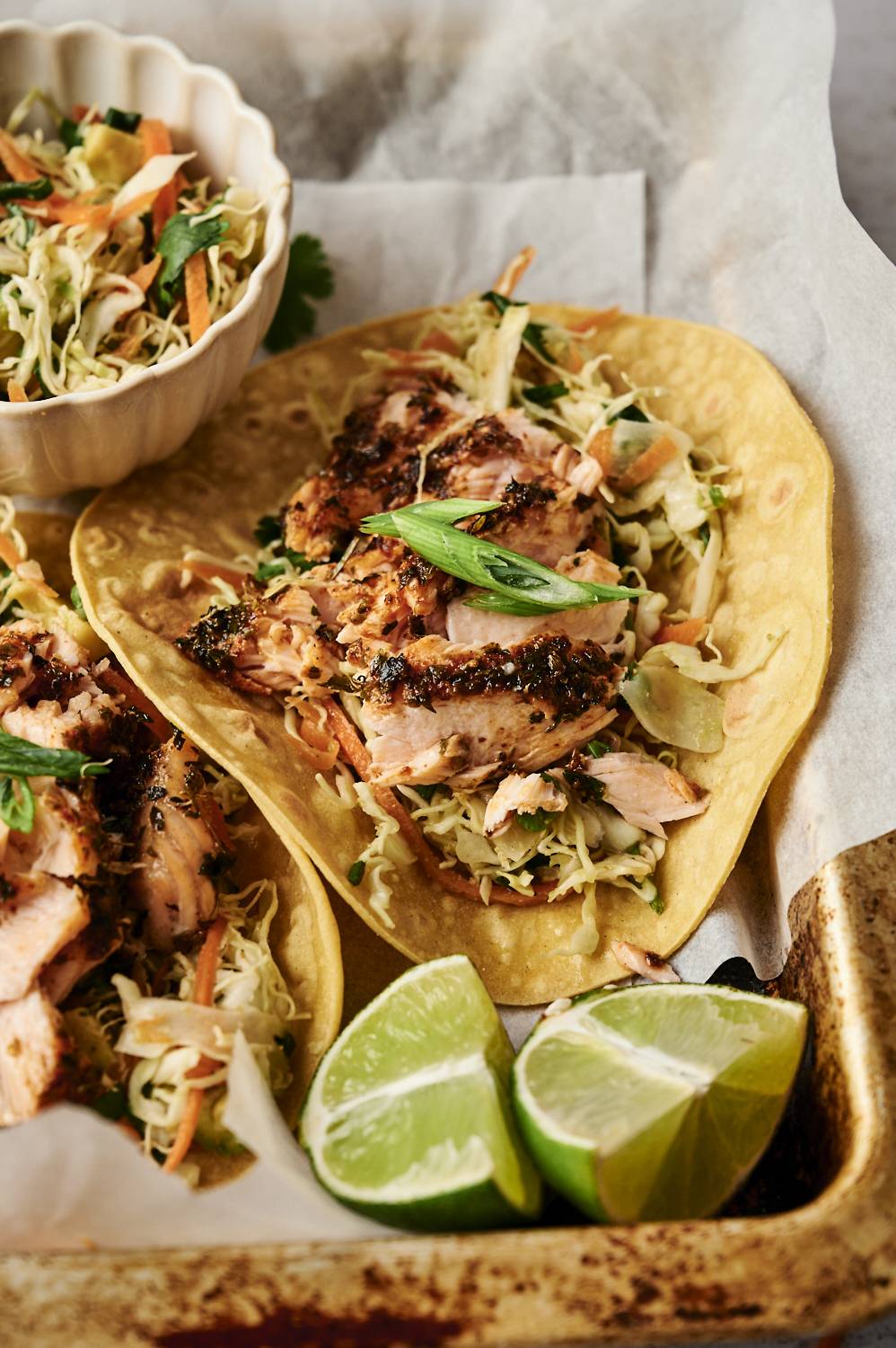 Salmon tacos are topped with slaw, green onions, and served with lime wedges on a tray lined with parchment paper.