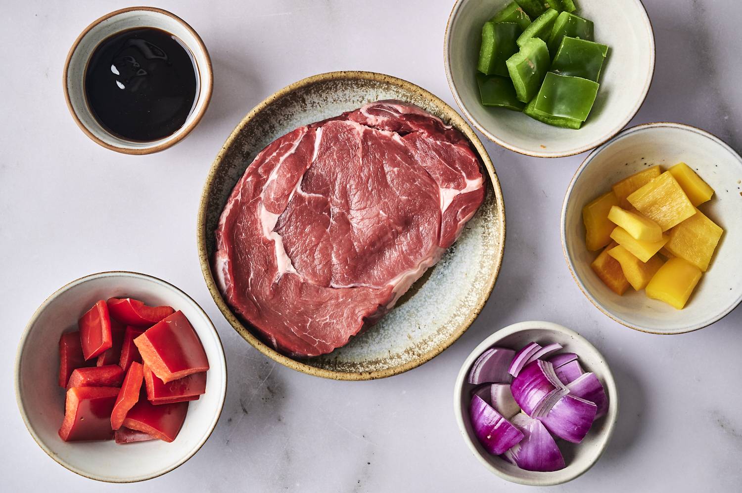 Raw rib-eye steak, surrounded by chopped red and green chili, yellow mango, red onions and a small bowl of sauce.