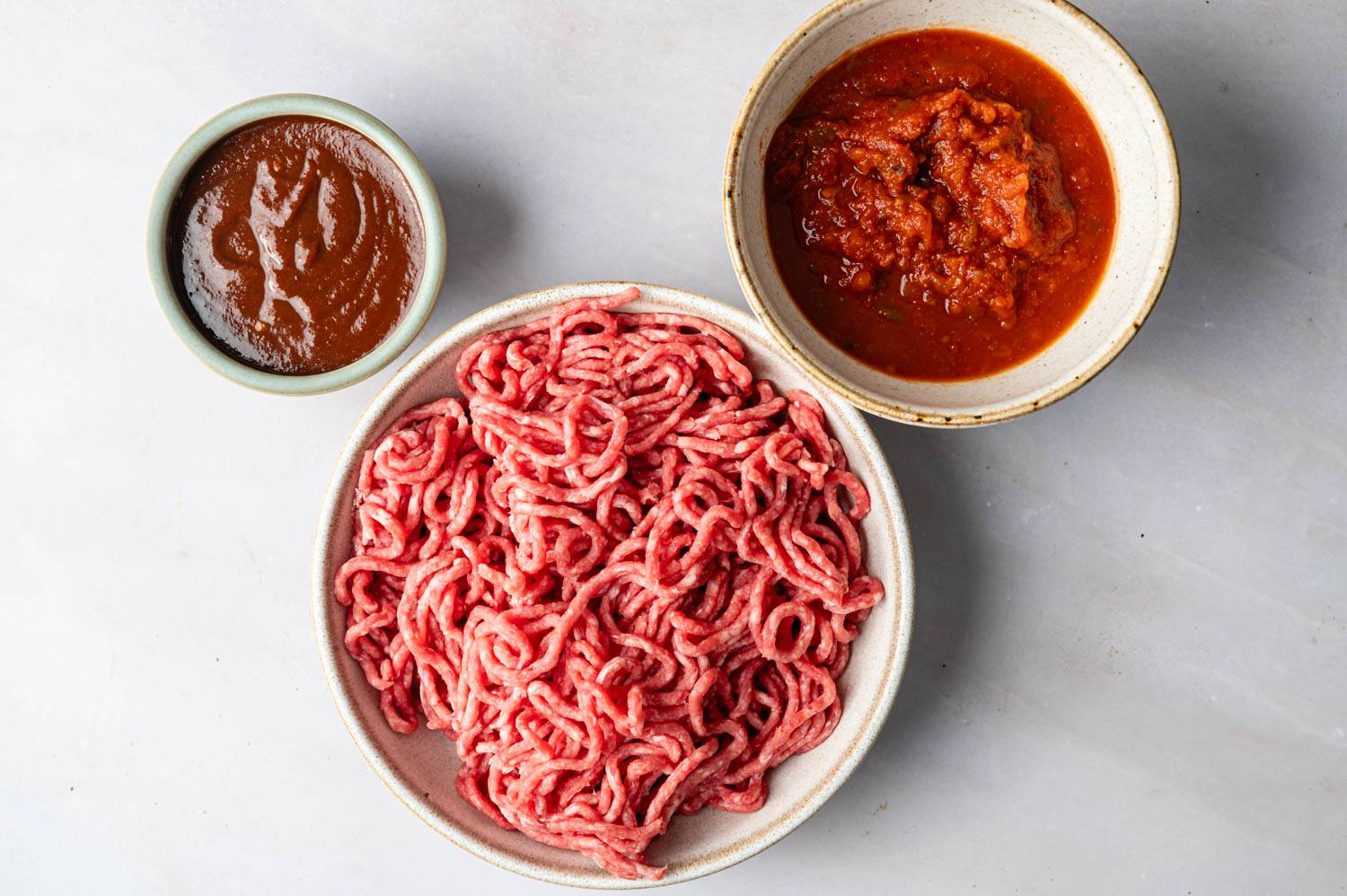Flat lay image of raw ground meat in large bowl and barbecue sauce and ketchup in small bowl on light gray surface.