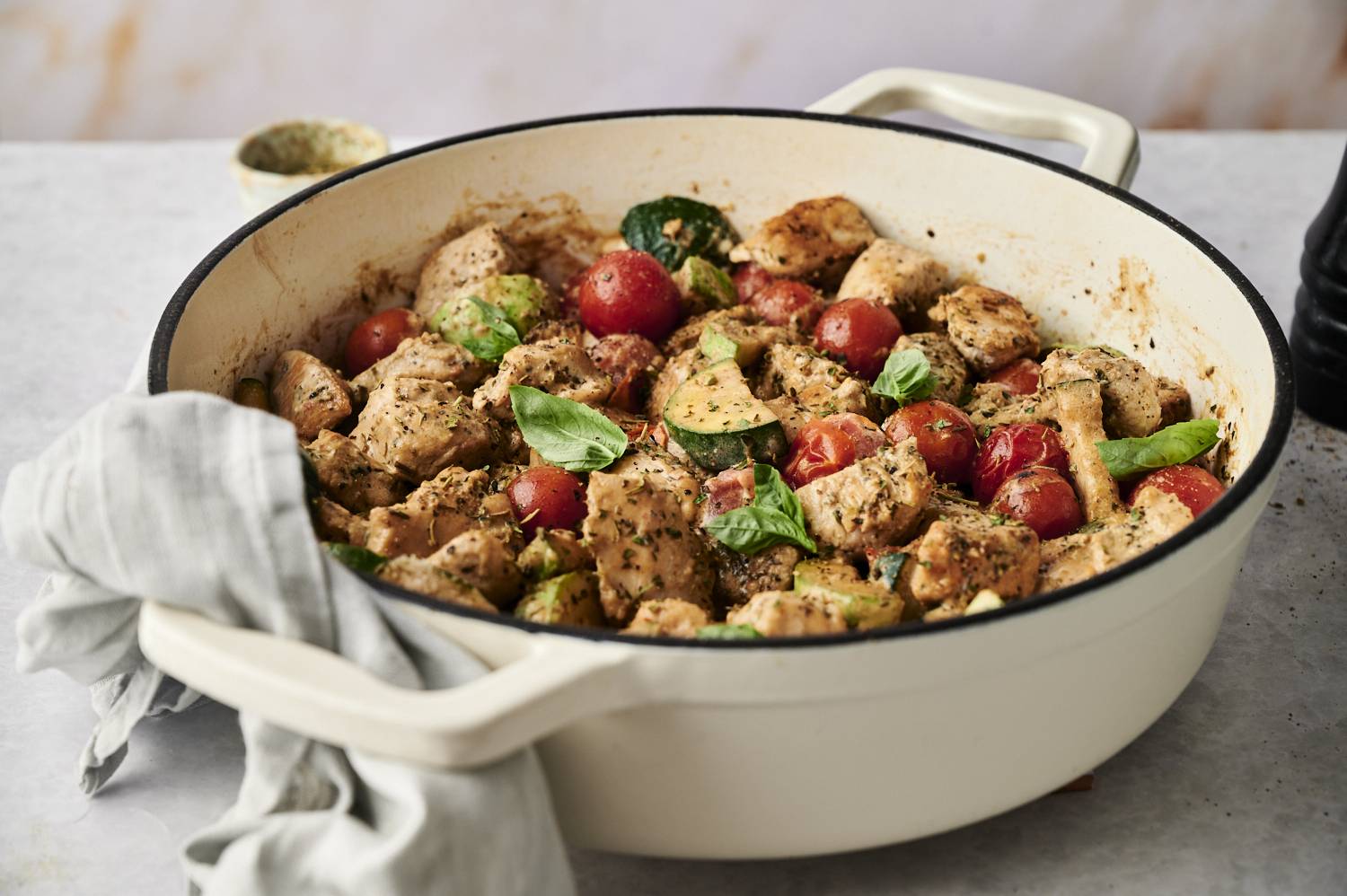 Creamy Tuscan chicken in a white Dutch oven filled with tender chicken, cherry tomatoes, zucchini and fresh basil, all coated with a savory herbal sauce. A gray kitchen towel draped over the handle.