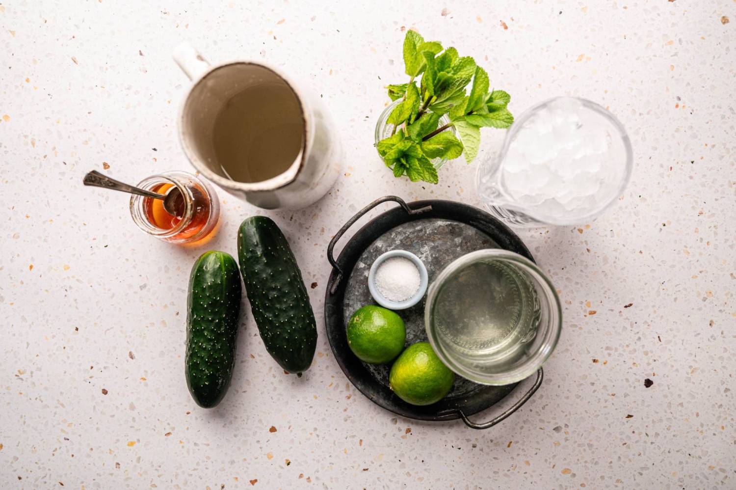 Ingredients for a refreshing drink arranged on a speckled surface: cucumber, honey, mint, ice, lime, water and salt on a small plate.
