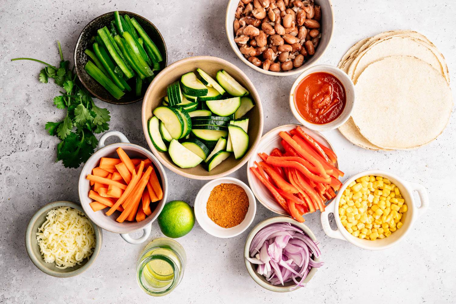 Assortment of fresh taco ingredients including sliced ​​zucchini, red peppers, corn, carrots, cheese, lime, beans, tortilla chips, salsa and spices on light surface.
