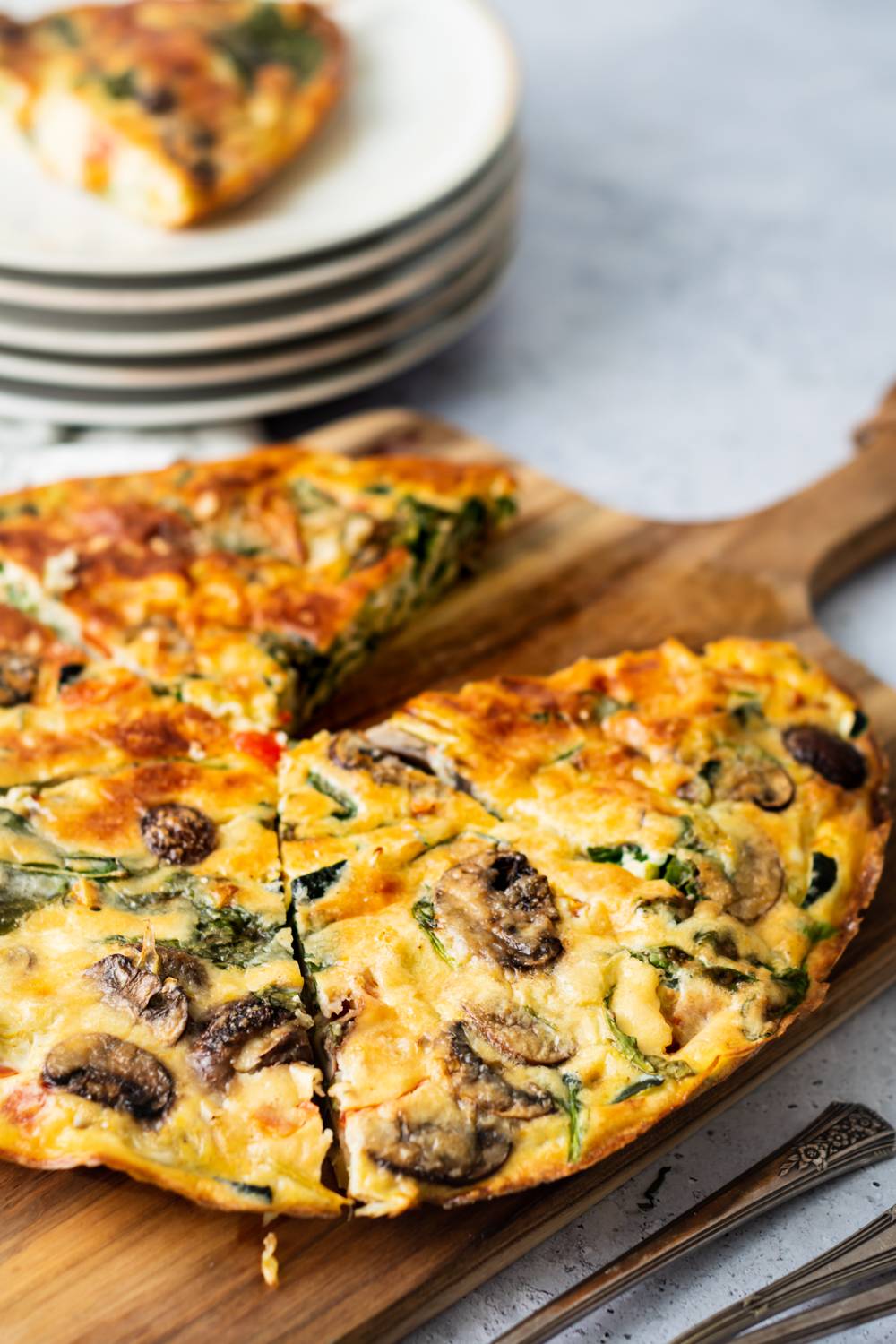 A golden, vegetable-filled frittata in a skillet with mushrooms, spinach, and peppers, served on a wooden board next to stacked plates and forks.