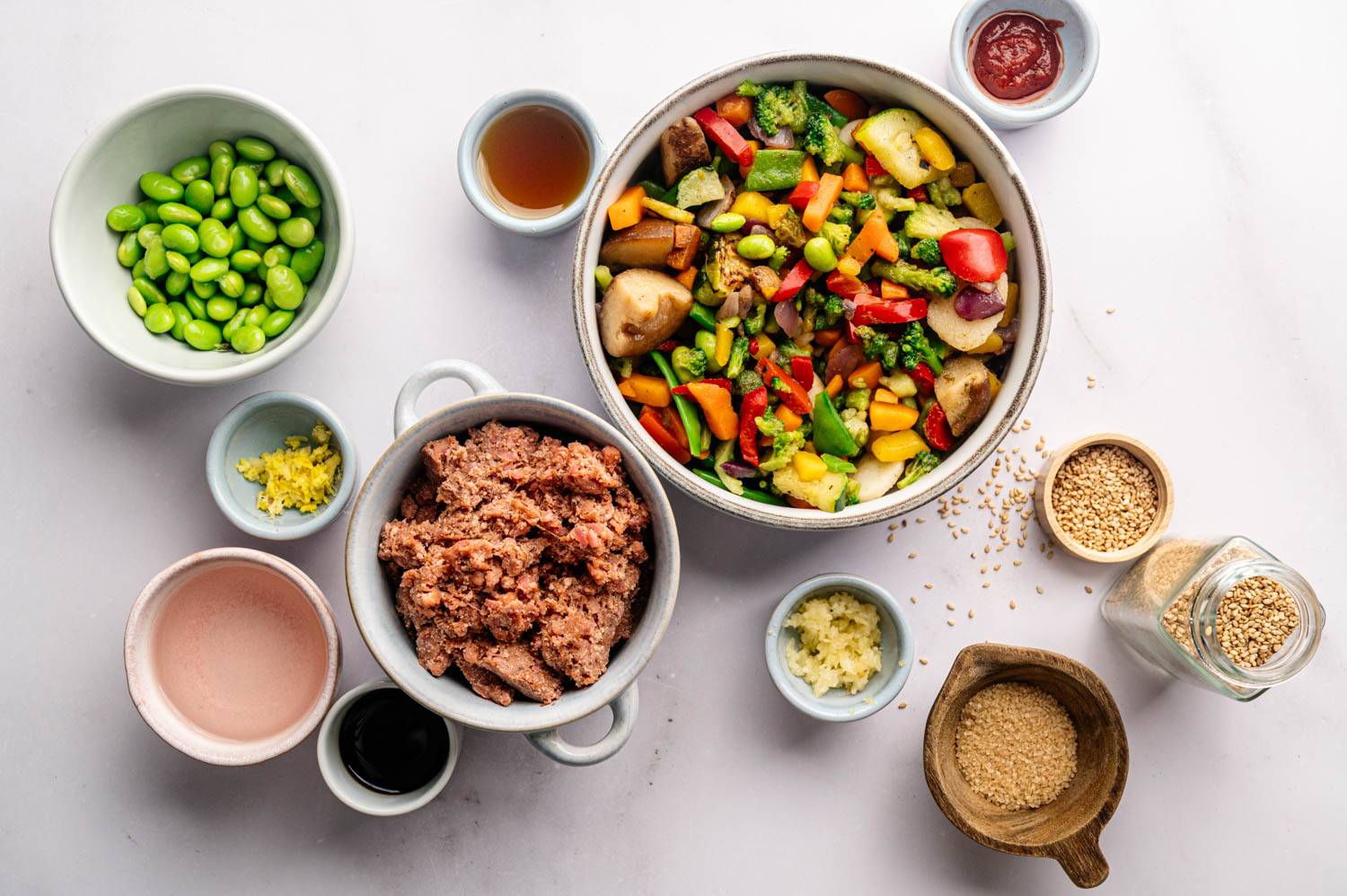 A colorful assortment of ingredients for a stir-fry, including chopped vegetables, ground meat, edamame, sauces, and spices, arranged on a white surface.