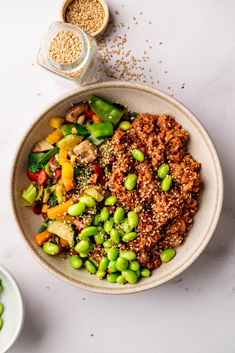 A bowl of vibrant vegan stir-fry with colorful vegetables, edamame, and quinoa, sprinkled with sesame seeds. A jar of sesame seeds is nearby.