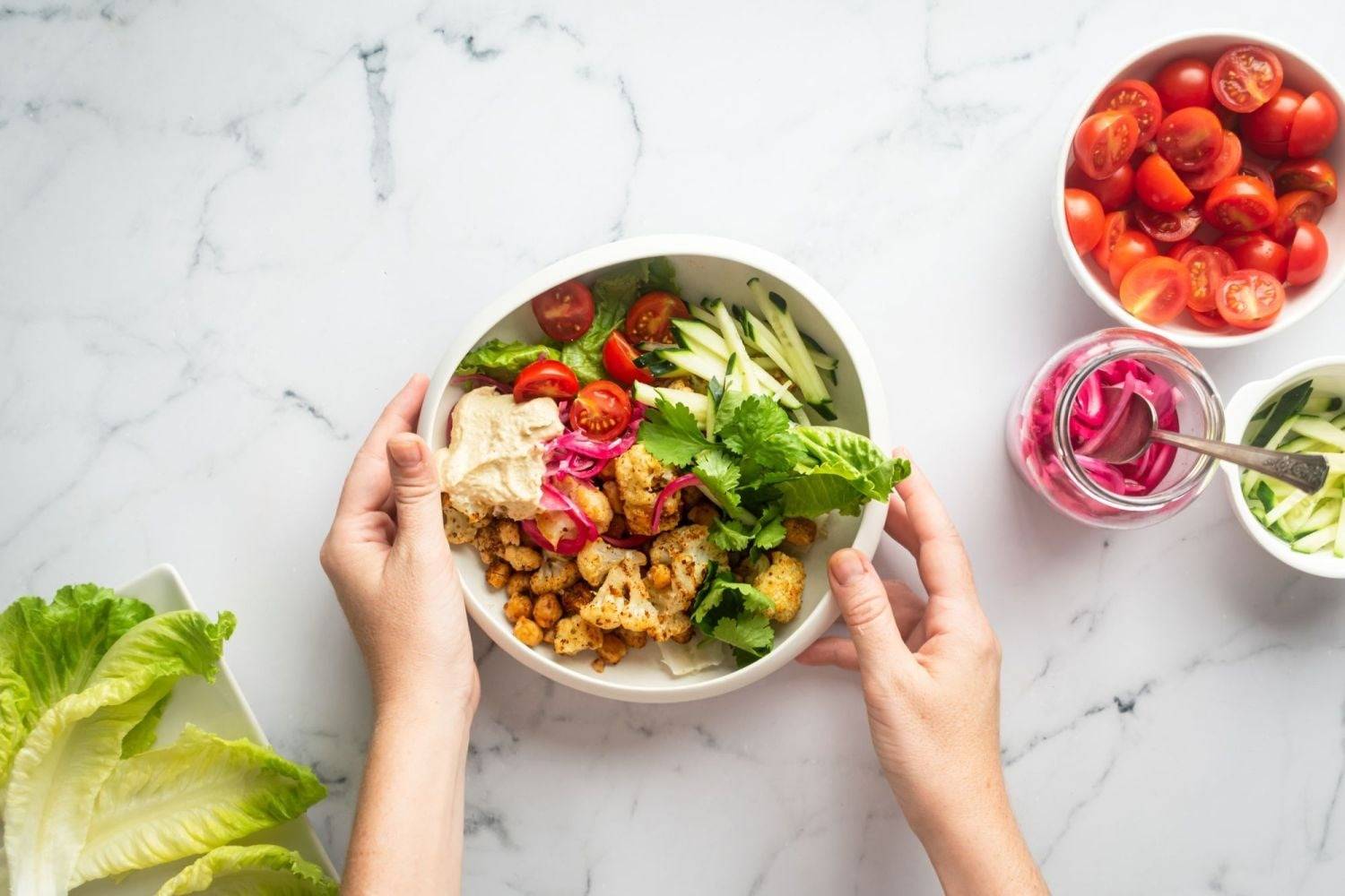 Cauliflower shawarma, chickpeas and vegetables in white bowls, held by two hands.