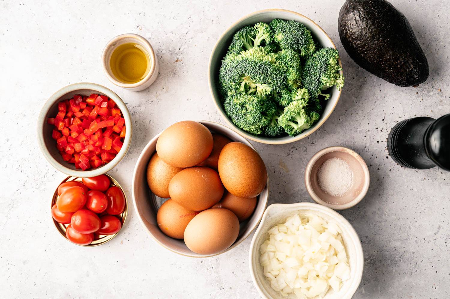 A variety of fresh ingredients on a light countertop: whole eggs, chopped onions, broccoli, diced red bell peppers, grape tomatoes, an avocado, olive oil, and salt.