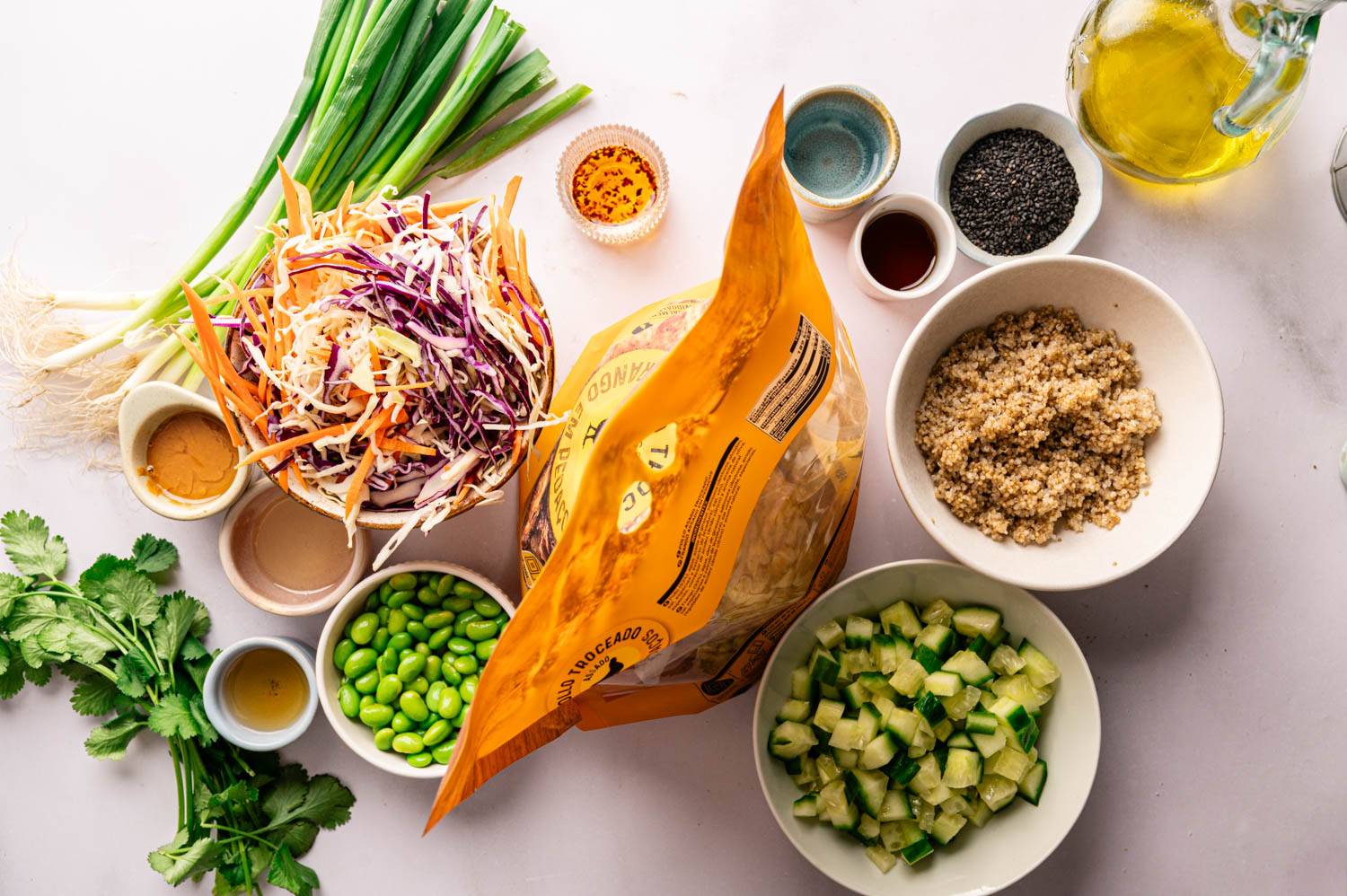 Fresh salad preparation scene with colorful salad, chopped cucumber, edamame, quinoa and dressing arranged on a white surface, evoking a vibrant healthy vibe.