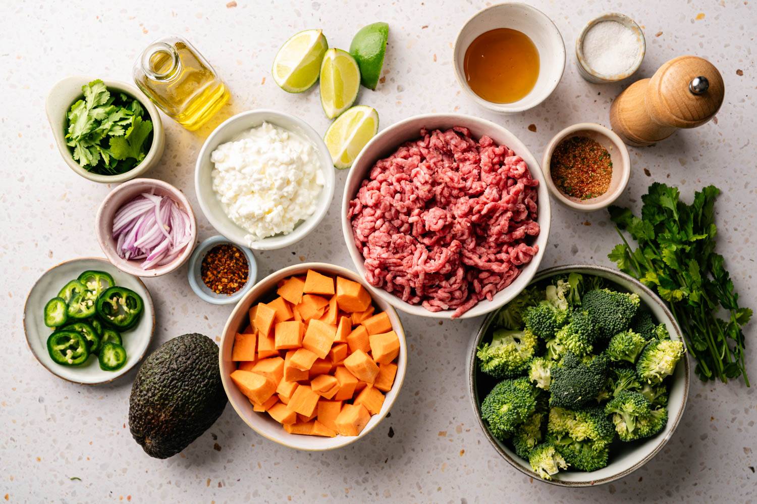 A variety of fresh ingredients are laid out on the light surface, including ground beef, broccoli, sweet potatoes, avocado, cilantro, lime, spices and oil.
