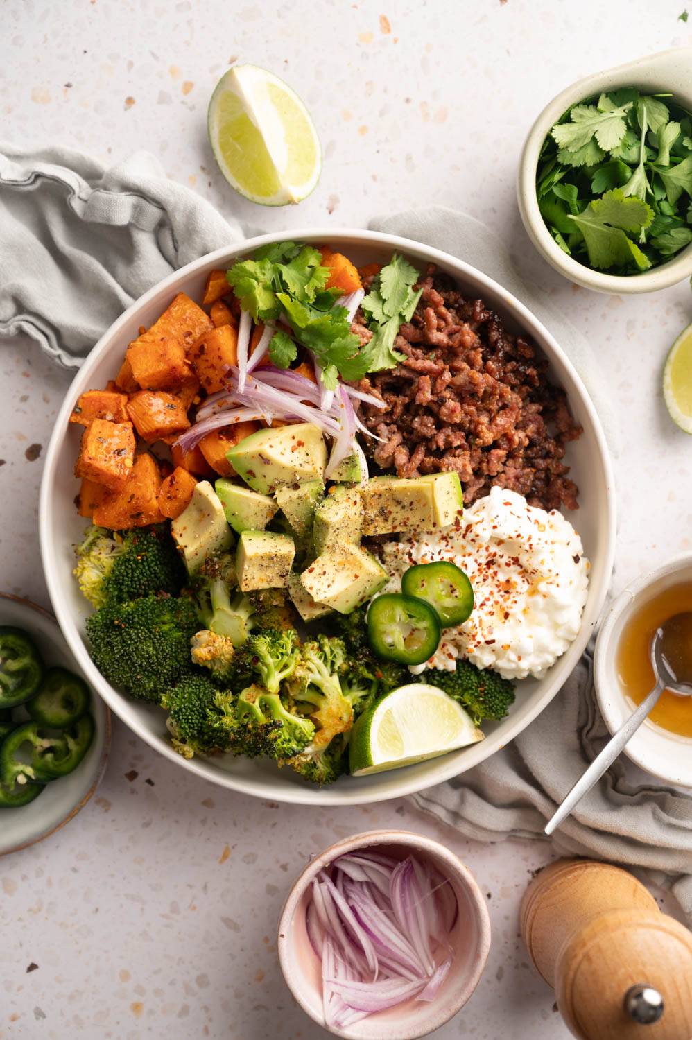 Colorful bowl with minced meat, roasted sweet potato, avocado, broccoli, jalapenos, onions, cilantro, mozzarella cheese and lime slices.