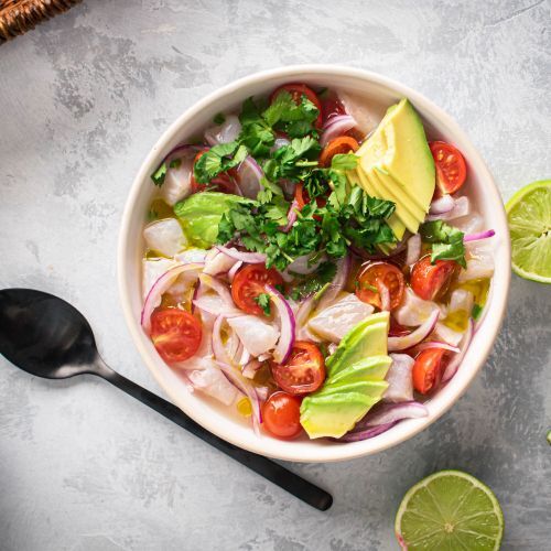 Ceviche with fish marinated in lime juice and served with cilantro, avocado, tomatoes, and red onion in a white bowl.