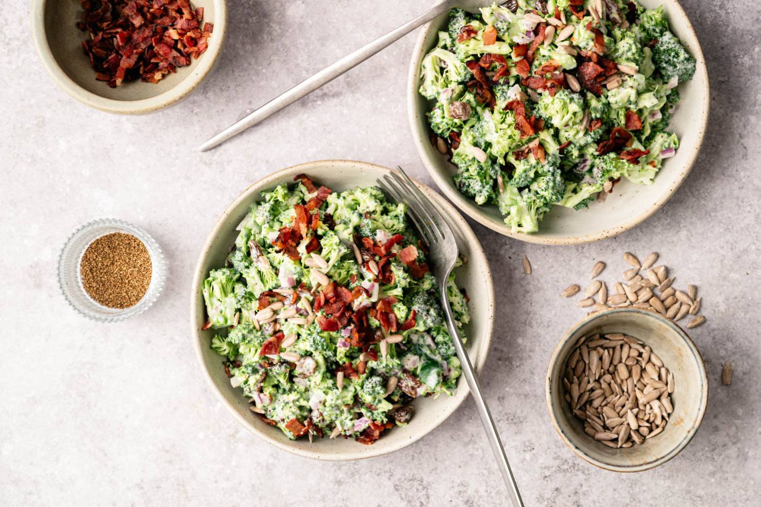 Two bowls of creamy broccoli salad topped with bacon and seeds are on a marble surface. Nearby are small bowls of bacon bits, sunflower seeds, and a fork.