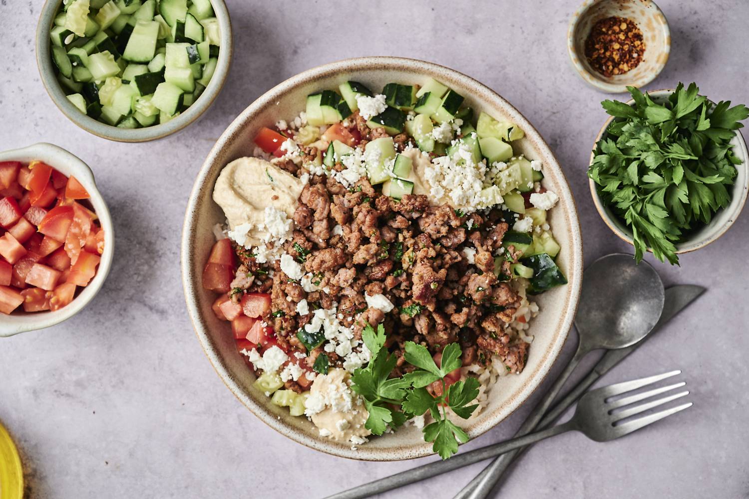 Chicken Kofta Bowls Bowl of hummus topped with seasoned ground meat, cucumber, tomatoes, feta, and fresh parsley, surrounded by ingredient bowls and utensils.