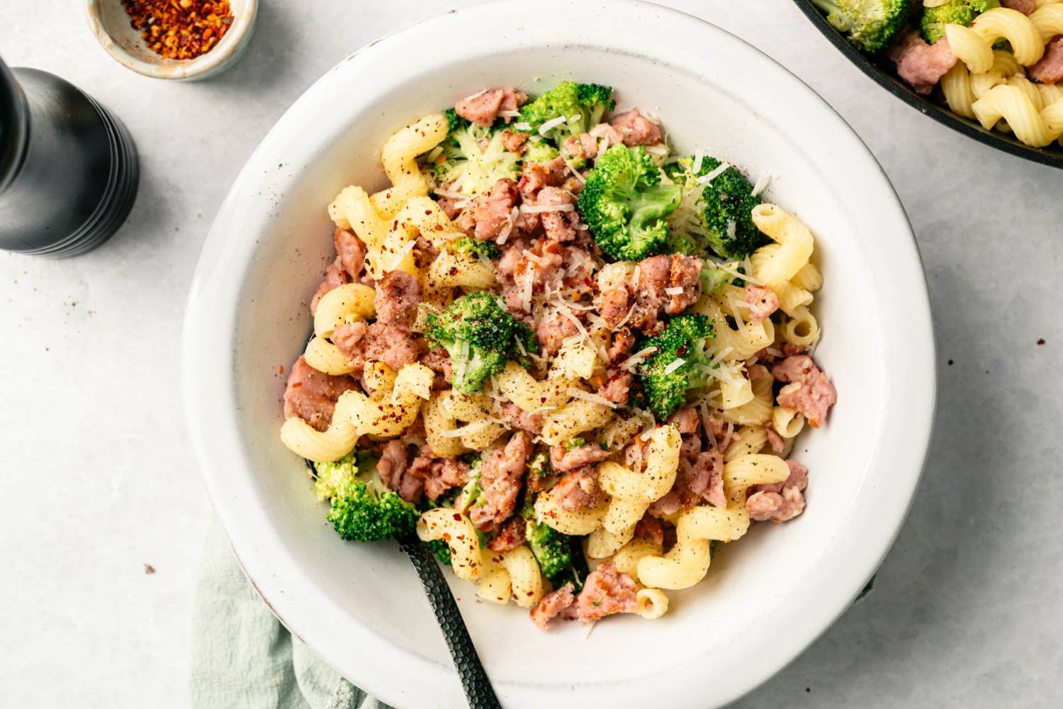 A bowl of curly cavatappi pasta mixed with broccoli and crumbled sausage, topped with grated cheese and black pepper. A black fork rests inside.