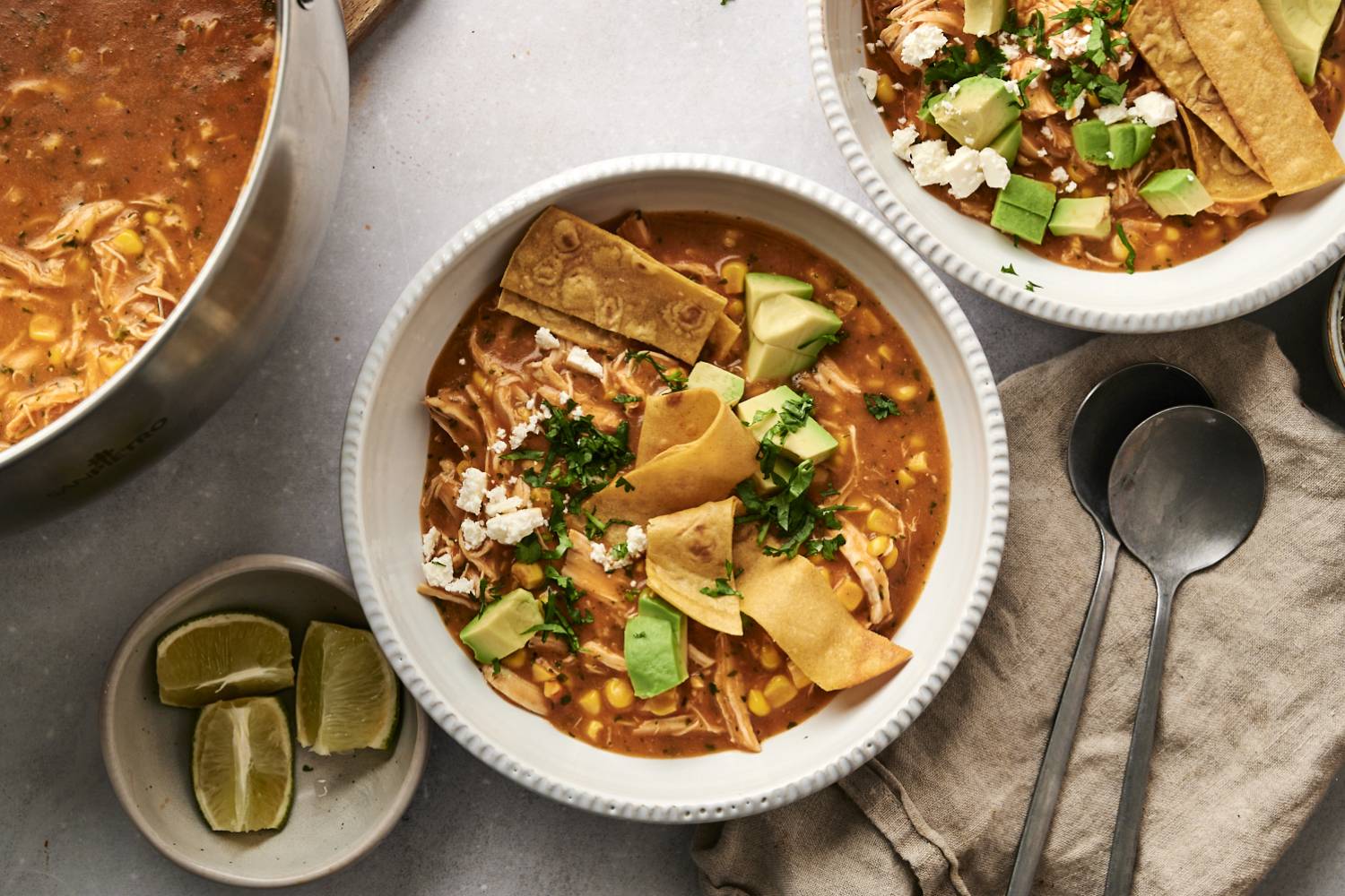 Chicken Tortilla Soup with Corn (Healthy & Freezer-Friendly!) Top-down view of chicken tortilla soup served in white bowls, garnished with crispy tortilla strips, avocado, cilantro, and crumbled cheese, with lime wedges and a pot of soup in the background.