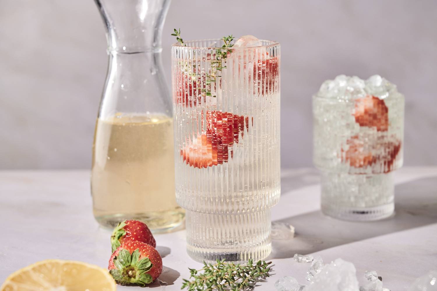 A tall glass of white wine spritzer garnished with strawberries and thyme sits next to a carafe of wine.