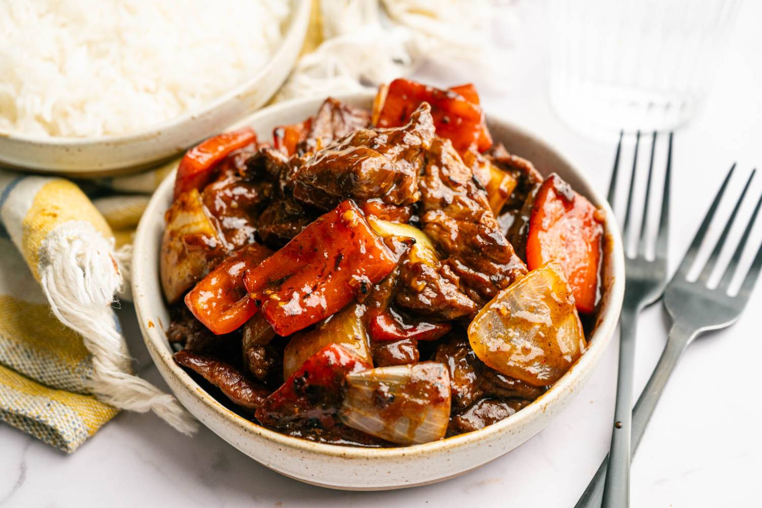 Copycat Beijing Beef A bowl of beef stir-fry with red peppers and onions in a savory sauce on a table next to a plate of white rice, a napkin, and forks.