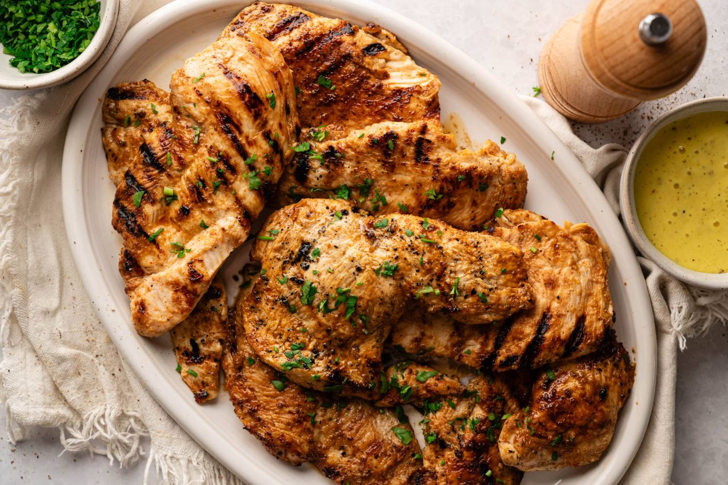 Grilled chicken breasts garnished with chopped parsley on a white plate. A small bowl of green herbs and a bowl of yellow sauce are nearby.