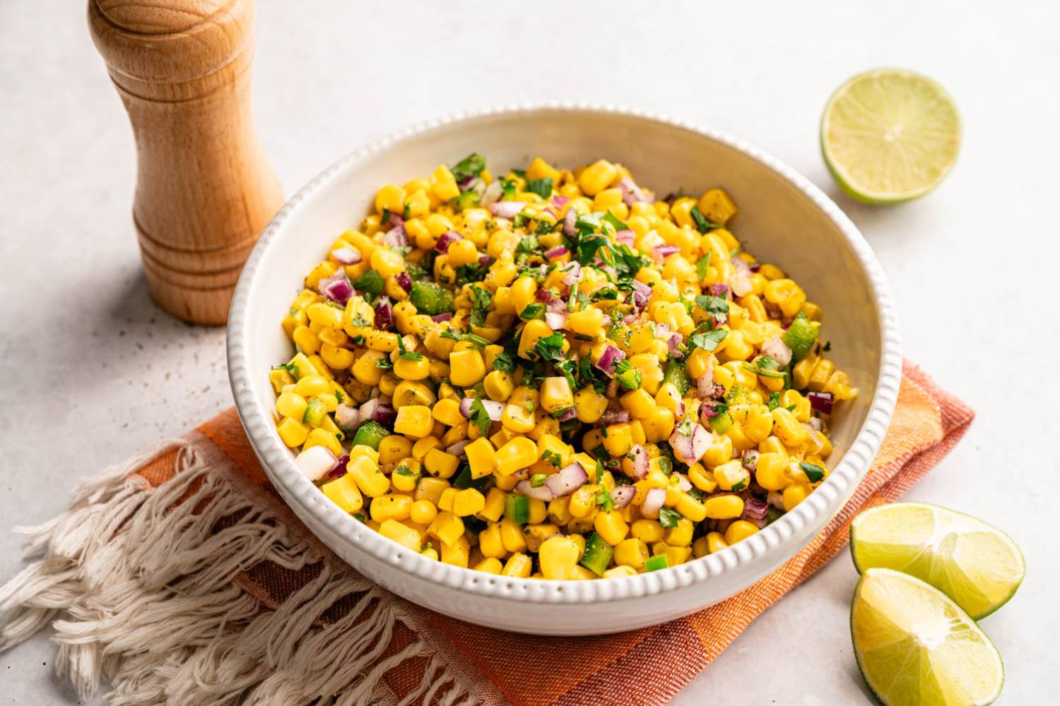 A vibrant corn salad with chopped red onions and cilantro in a white bowl, set on a fringed orange cloth. Lime slices and a pepper grinder nearby.