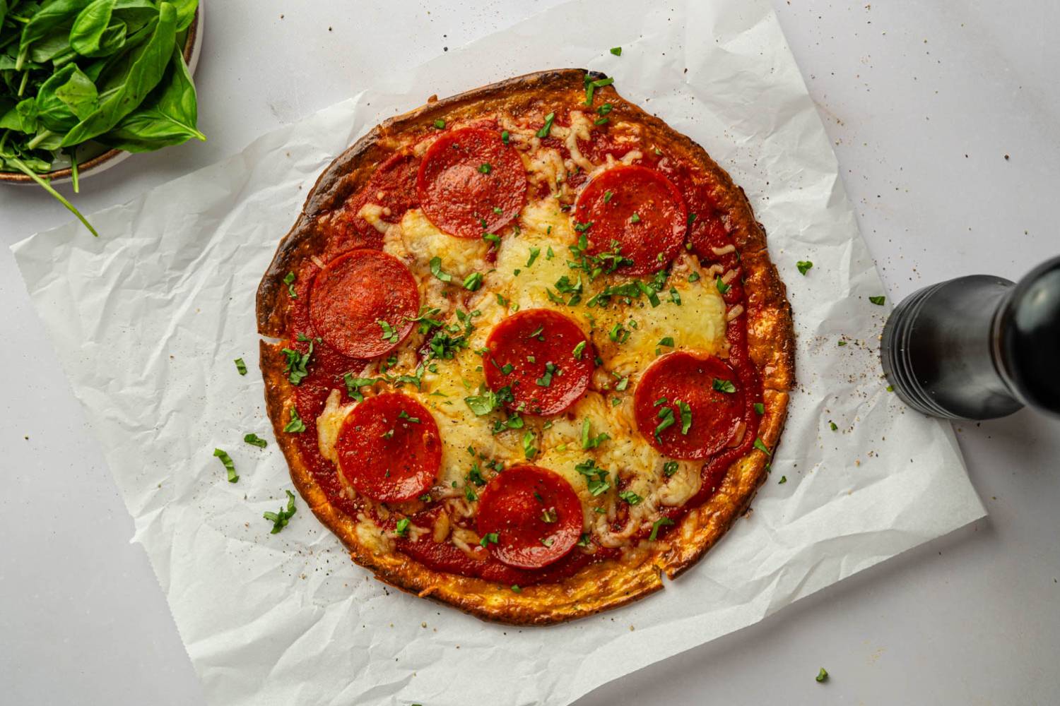 Whole cottage cheese pepperoni pizza with melted mozzarella cheese, turkey pepperoni, and chopped basil on parchment paper, viewed from above.