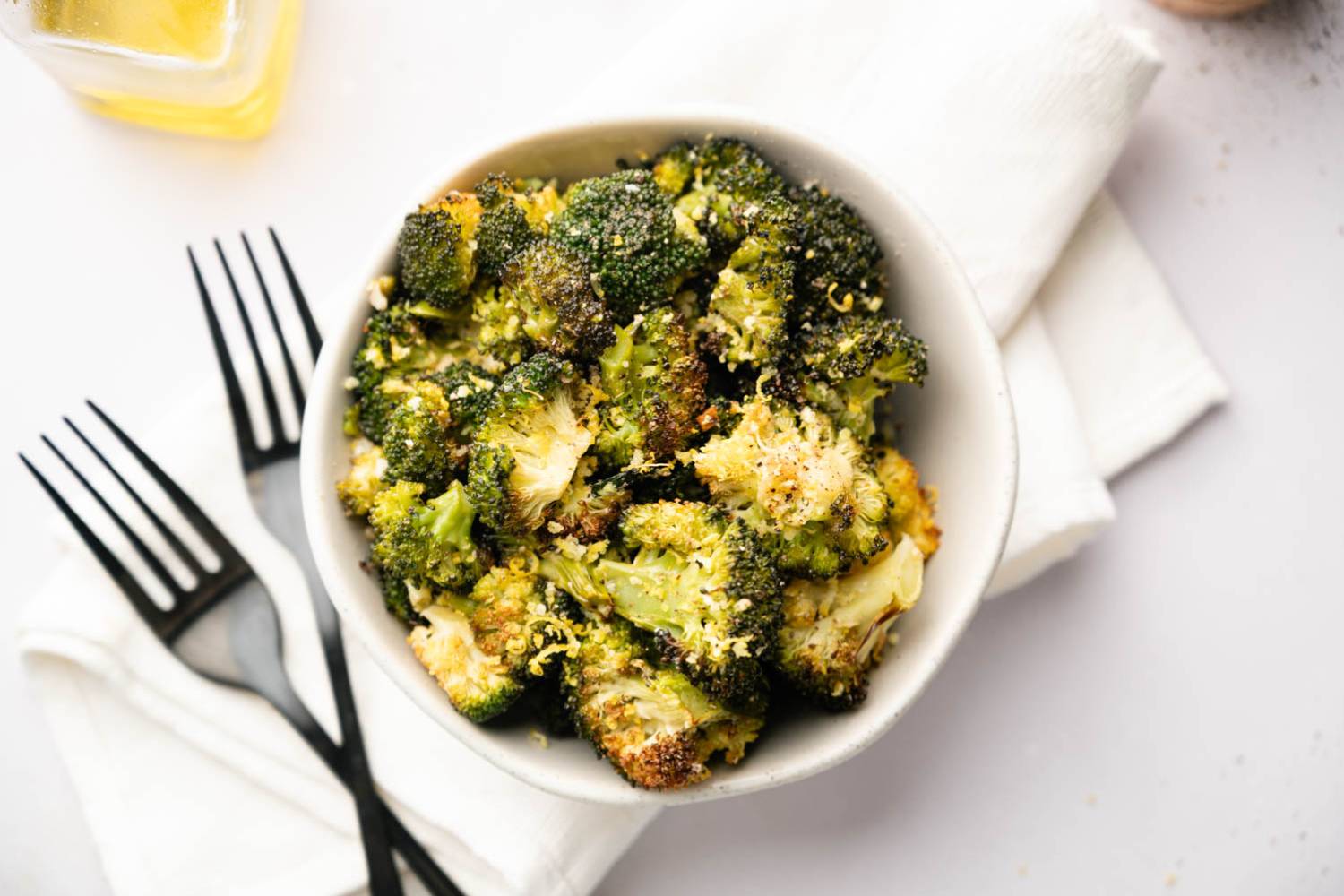 Crispy Baked Broccoli Bowl of roasted broccoli topped with grated cheese, set on white napkin. Two black forks lie beside, with a glass of yellow liquid nearby.