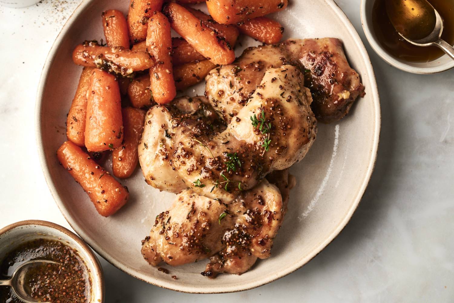 Overhead shot of bone-in chicken thighs coated in a grainy honey mustard glaze and fresh thyme, plated alongside roasted whole baby carrots on a cream-colored ceramic dish, with small bowls of pan sauce and honey positioned at the edges of the frame.