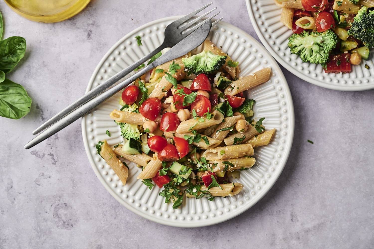 Easy Vegetable Pasta Salad A vibrant plate of pasta mixed with cherry tomatoes, broccoli, and herbs, accompanied by a glass of olive oil and fresh basil leaves.