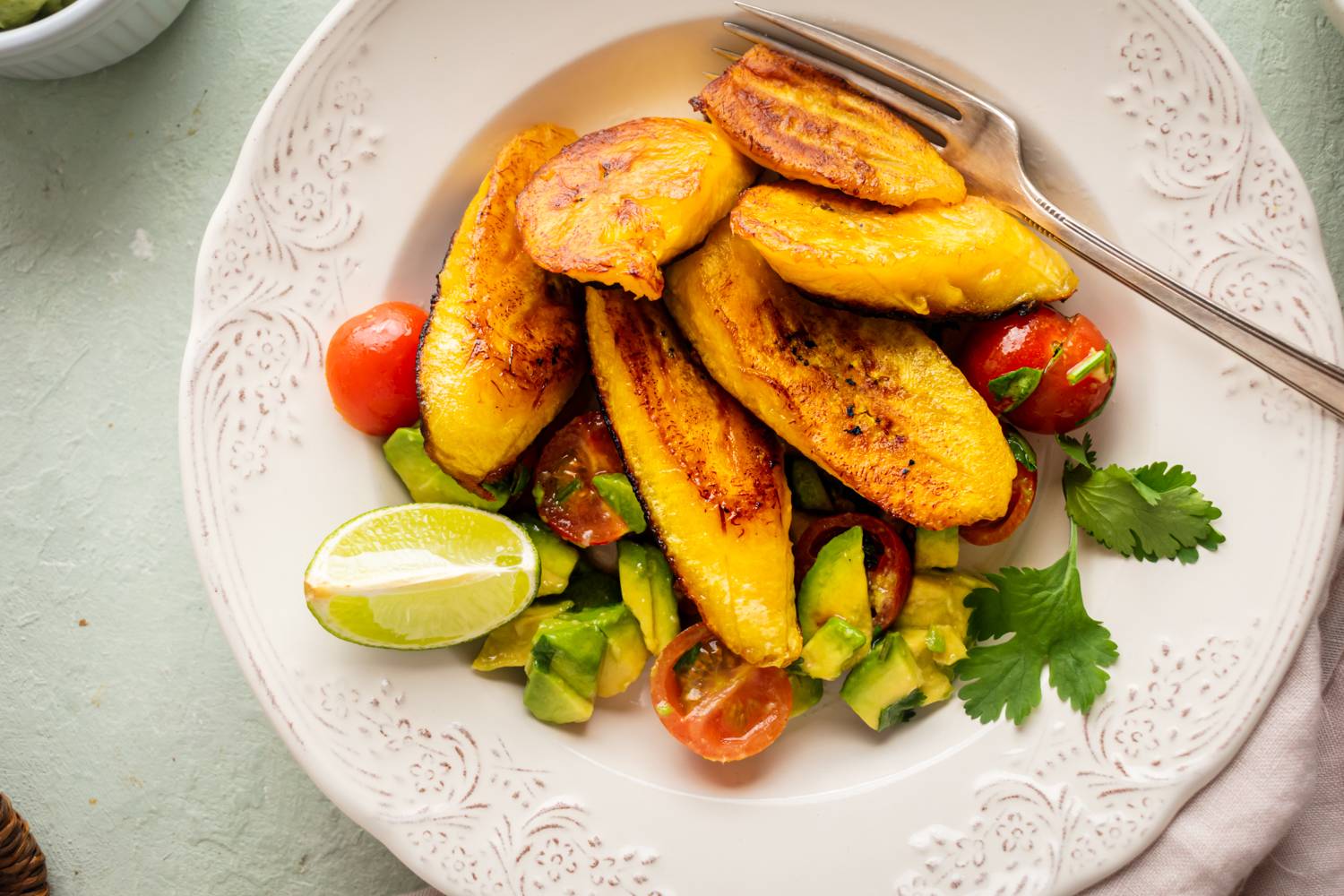 Crispy Fried Plantains A white plate with golden-brown fried plantains, sliced avocado, cherry tomatoes, lime wedge, and cilantro. The setting is light, fresh, and inviting.