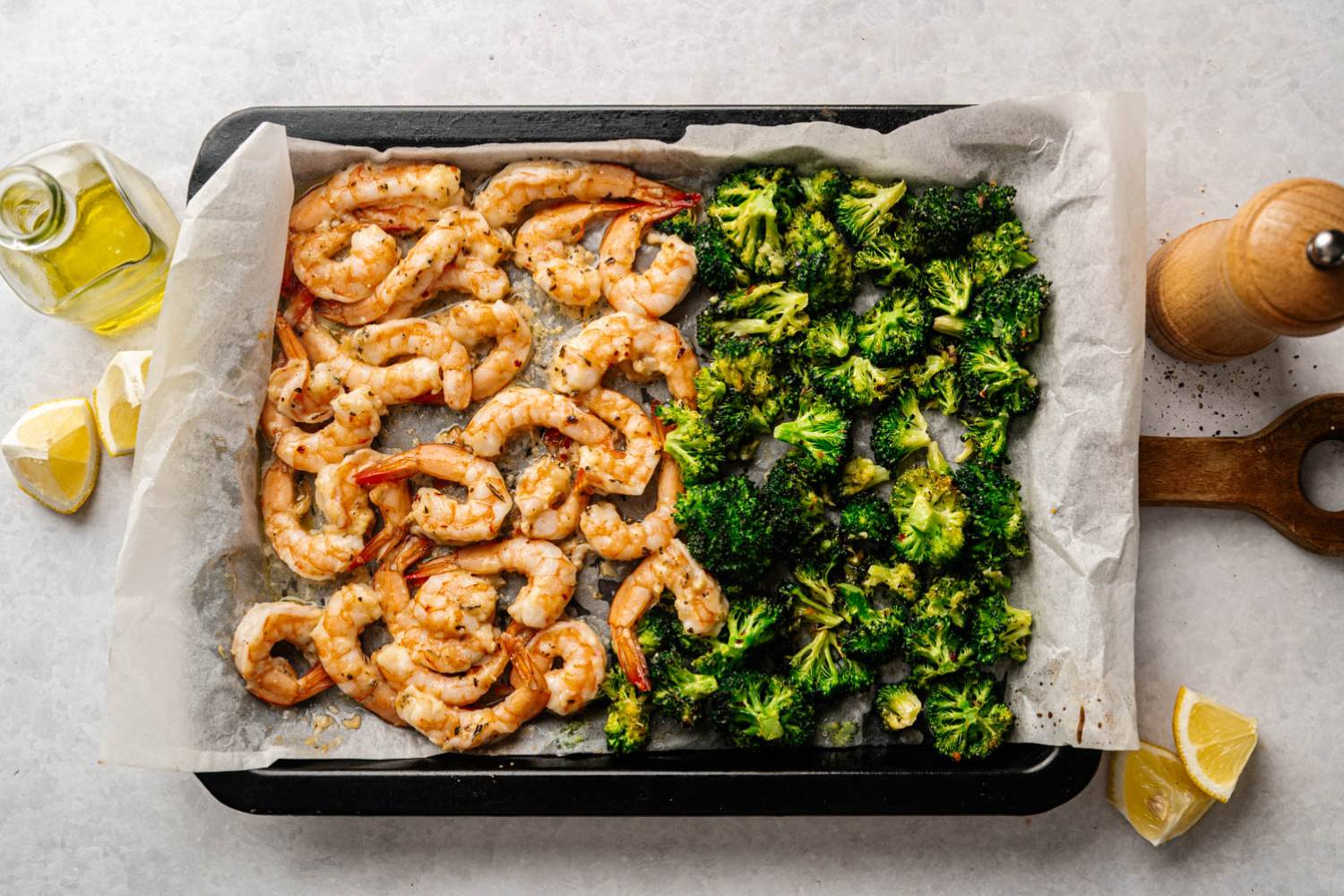 Garlic Parmesan Roasted Shrimp and Broccoli Roasted shrimp and broccoli on a baking sheet lined with parchment paper. Olive oil, lemon wedges, and a pepper mill are nearby. Bright and appetizing.