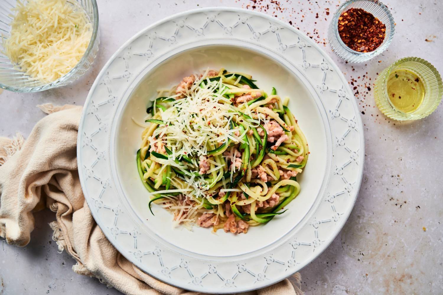 20 Lightened-Up Pasta Alternatives Garlic Parmesan zucchini noodles served in a white dish, topped with grated cheese and red pepper flakes, accompanied by ingredients like olive oil and shredded Parmesan cheese.