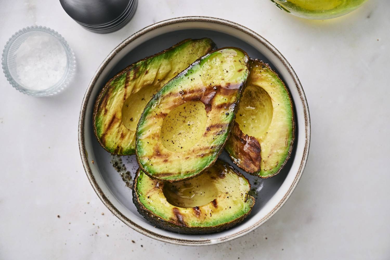 Grilled Avocados Grilled avocado halves drizzled with oil and sprinkled with pepper, served in a bowl beside coarse salt.
