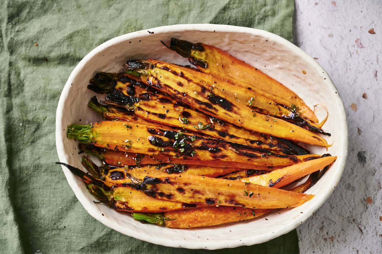 Easy Grilled Carrots A white bowl filled with charred orange carrots topped with herbs, resting on a green cloth and surrounded by a textured surface.