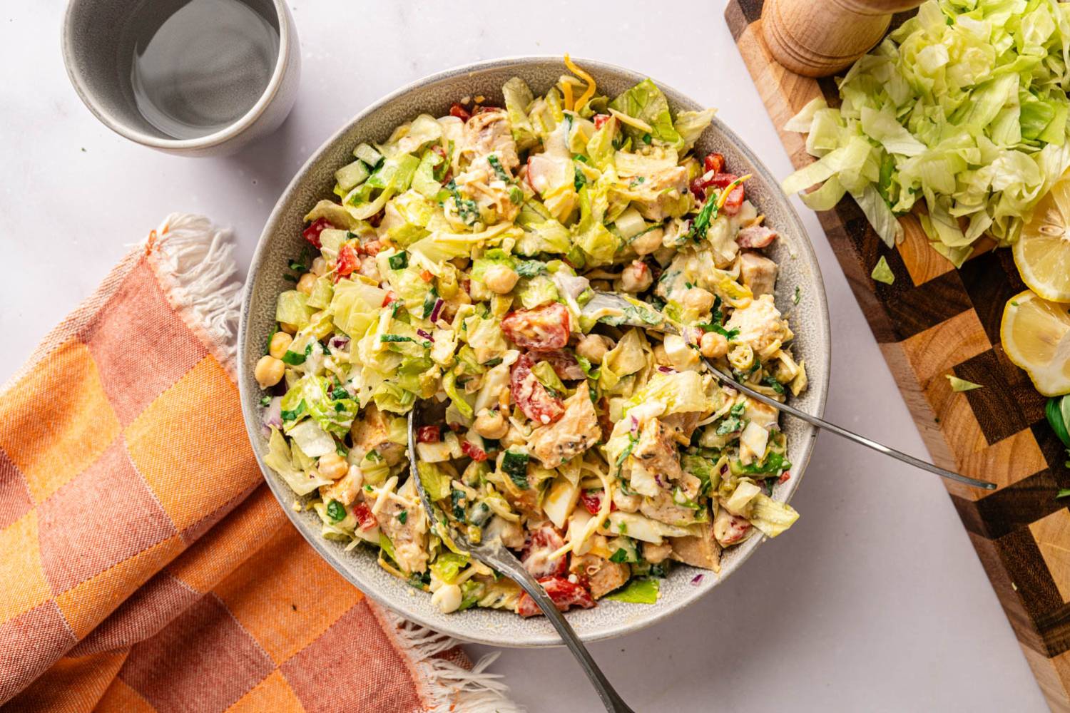 Grilled Chicken Chopped Salad Overhead view of grilled chicken chopped salad in a stoneware bowl with two forks resting inside, surrounded by an orange checkered napkin, a wooden pepper grinder, sliced lemons, and shredded lettuce on a cutting board.
