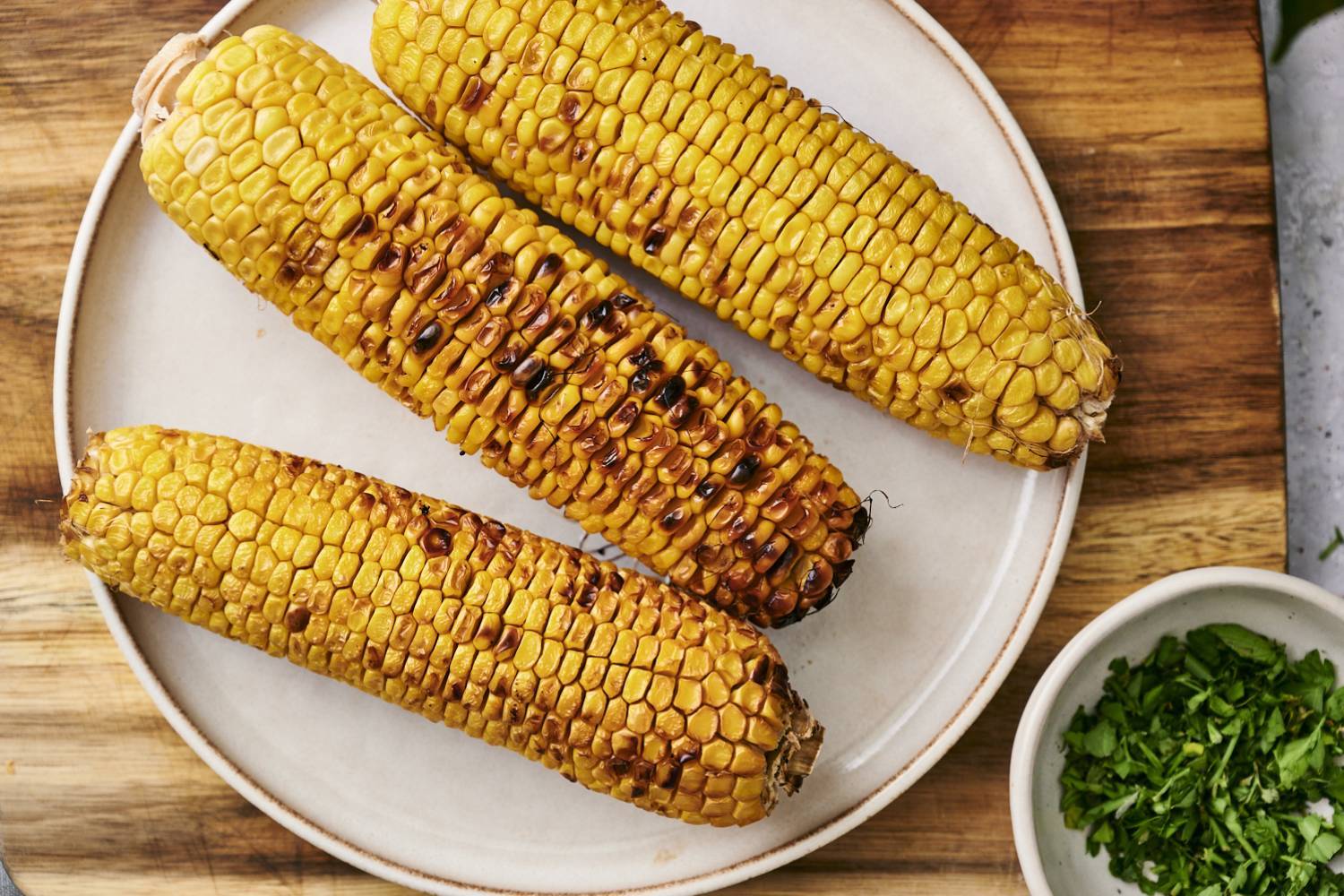 Healthy Grilled Corn Ideas Three ears of grilled corn with lightly charred kernels arranged on a white plate, with a small bowl of chopped fresh cilantro on a wooden cutting board.