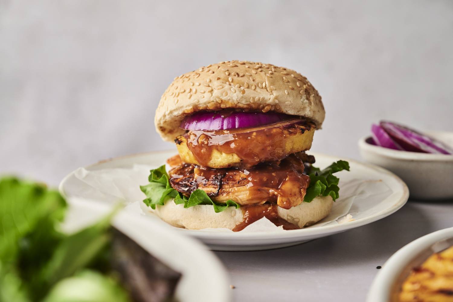 Grilled Hawaiian Chicken Sandwiches A delicious sesame seed burger with grilled chicken, lettuce, a slice of pineapple, red onion, and a savory sauce on a white plate.