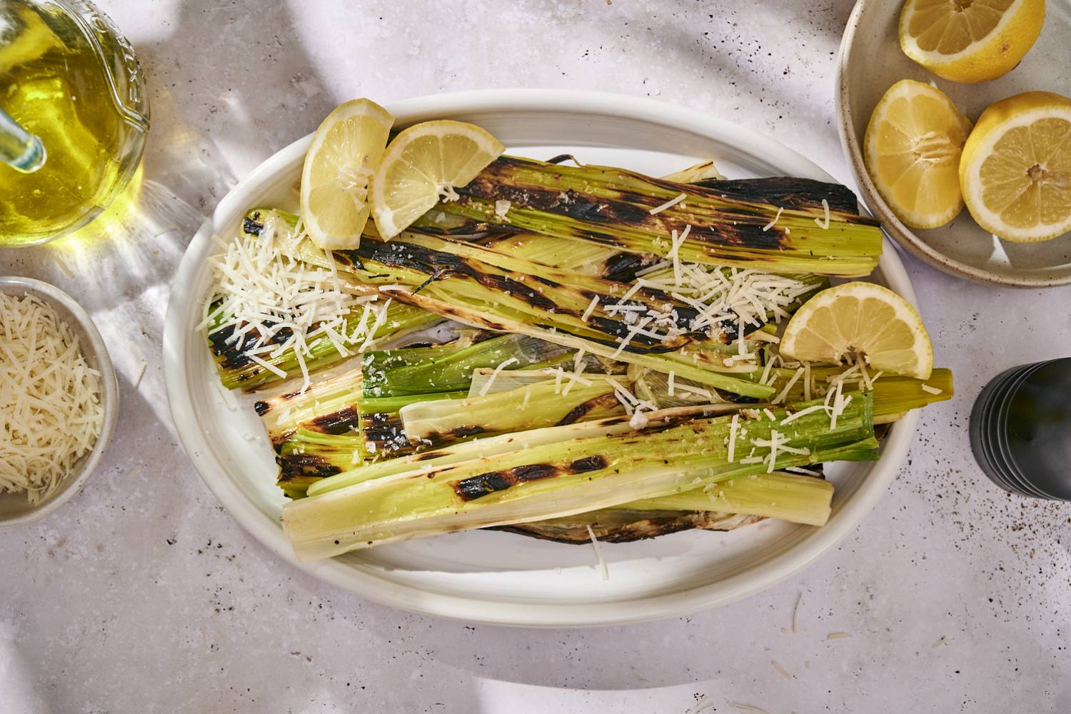 Grilled Leeks Charred grilled leeks served on a white platter with lemon wedges and a sprinkle of shredded Parmesan cheese.