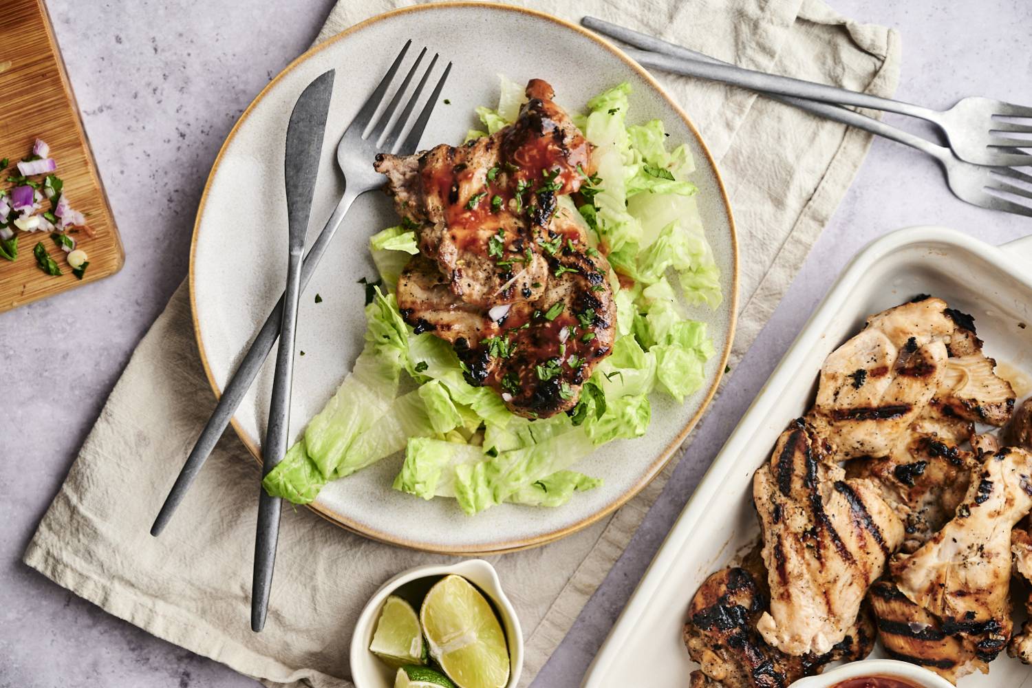 Grilled Sriracha Barbecue Chicken Grilled chicken topped with sauce on a bed of lettuce, served with lime wedges and garnished with cilantro, next to a platter of more chicken.