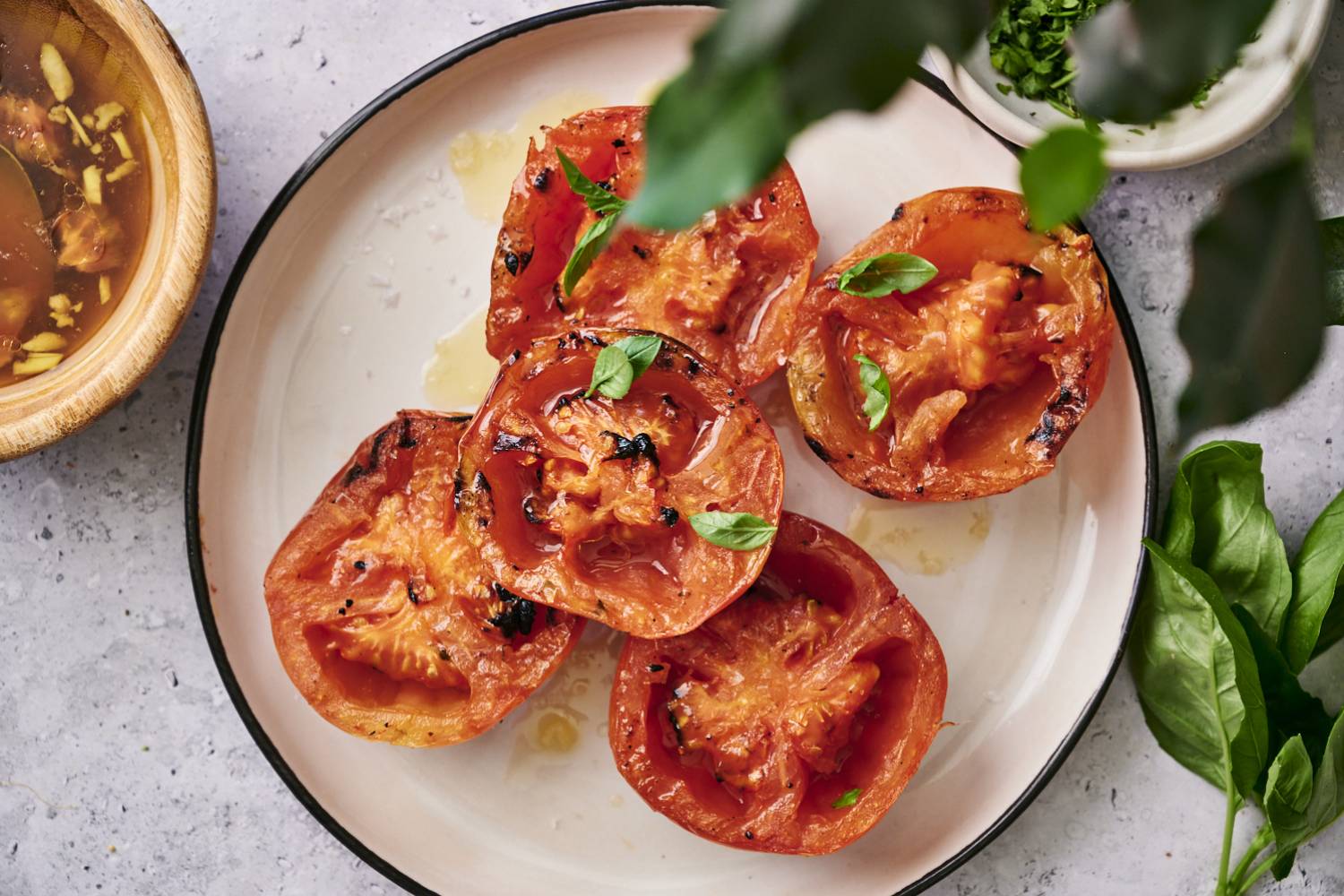 Grilled Tomatoes Grilled tomatoes topped with fresh basil, drizzled with olive oil, served on a round plate alongside a bowl of broth.